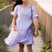 Woman in a light purple dress walking on a wooden boardwalk by the beach.