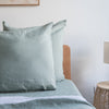 Harbour Sage European linen pillowcaseson a bed with a wooden headboard and lamp in the background.
