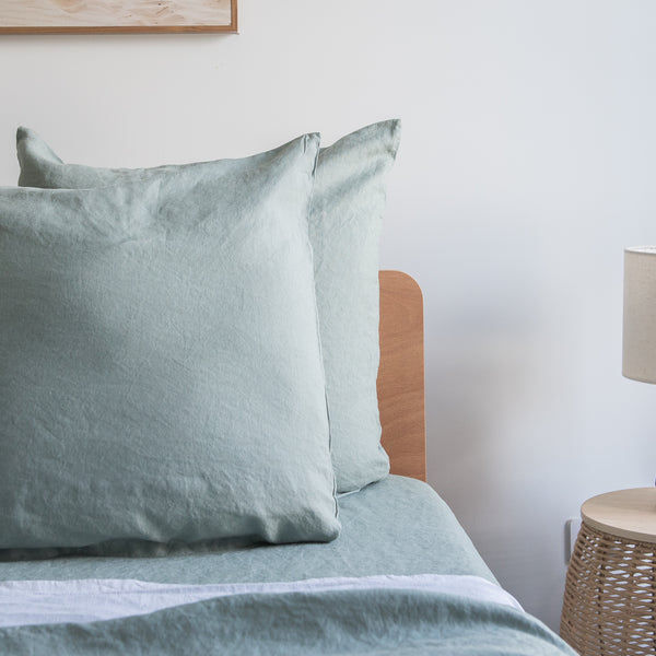 Harbour Sage European linen pillowcaseson a bed with a wooden headboard and lamp in the background.