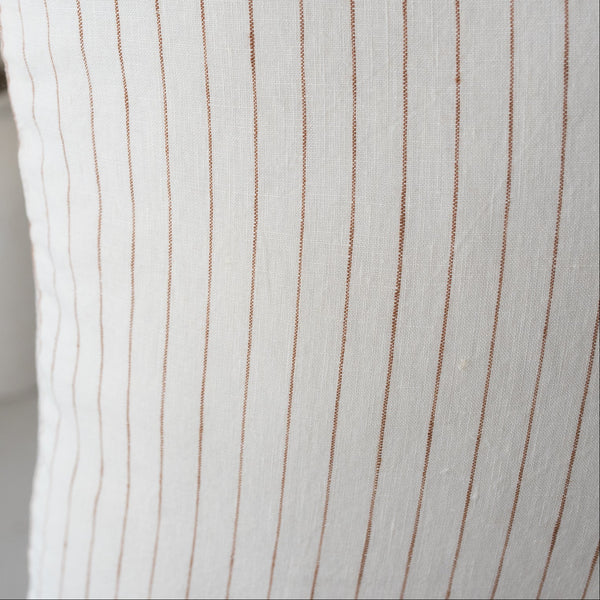 Close-up of a white pillow with brown stripes and a blurred vase in the background.