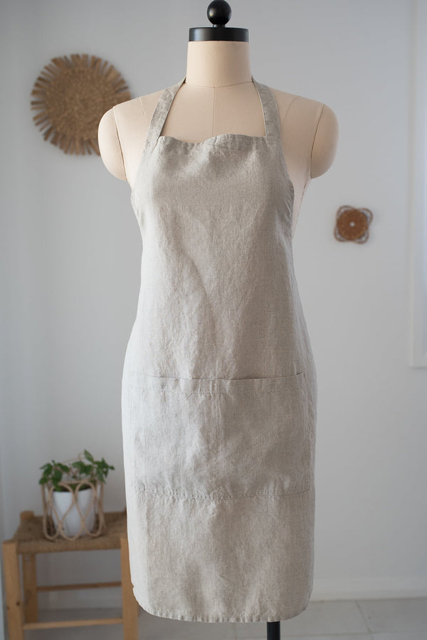 Beige apron on a mannequin against a white wall with a small plant and wooden stool in the background.